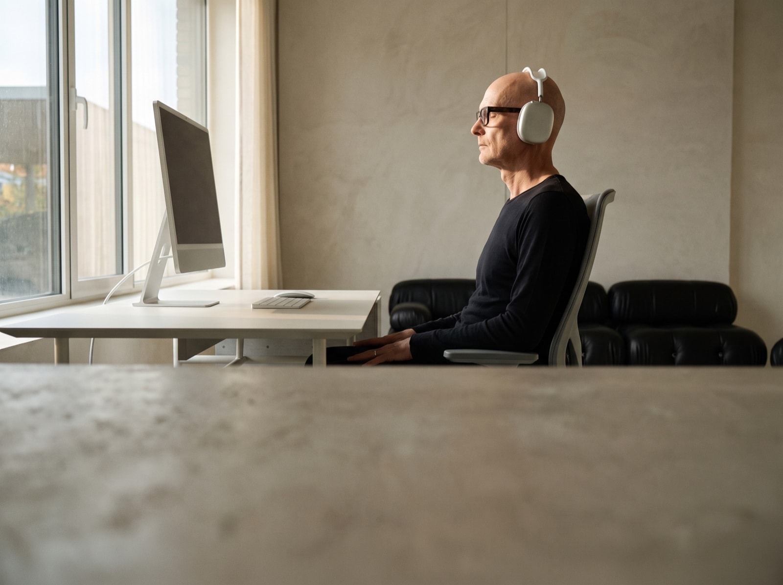 A man in headphones sits at a desk by a window, eyes closed, the monitor dark.