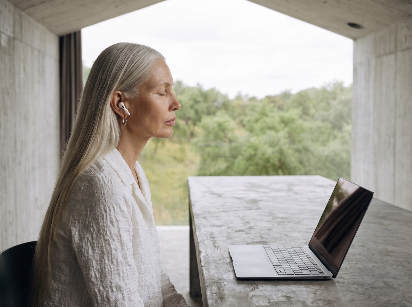 A woman with white hair sits at a concrete table with a laptop, eyes closed, trees beyond the open wall.