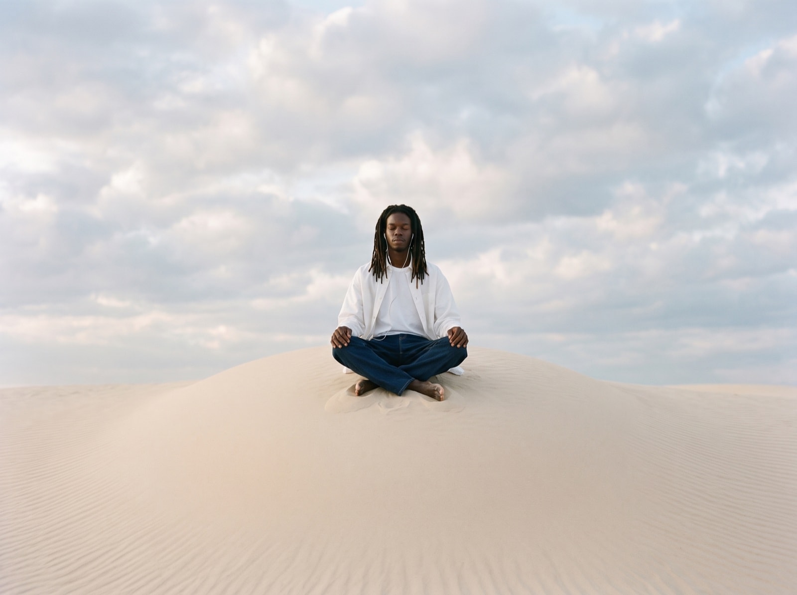 A man sits cross-legged on the crest of a sand dune under a wide cloudy sky.