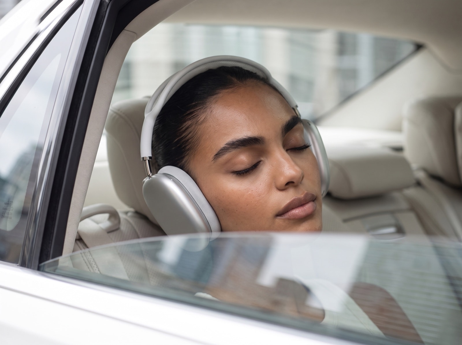 A woman rests her head against the back seat of a car with headphones on, eyes closed, the city soft behind the window.