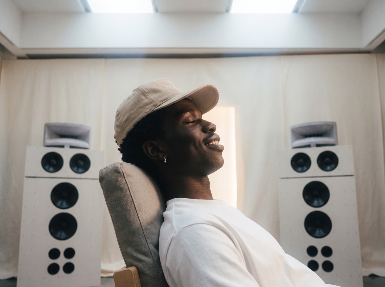 A man in a cap leans back in a chair between two tall speakers, smiling with his eyes closed.