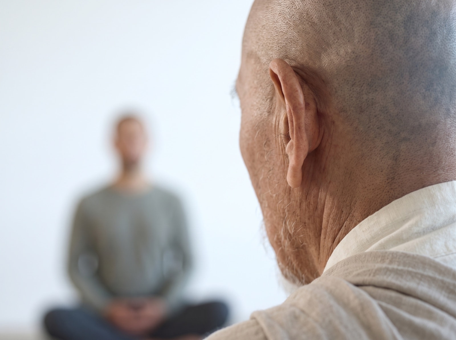 An elder watches from close by as a student sits cross-legged in a white room.
