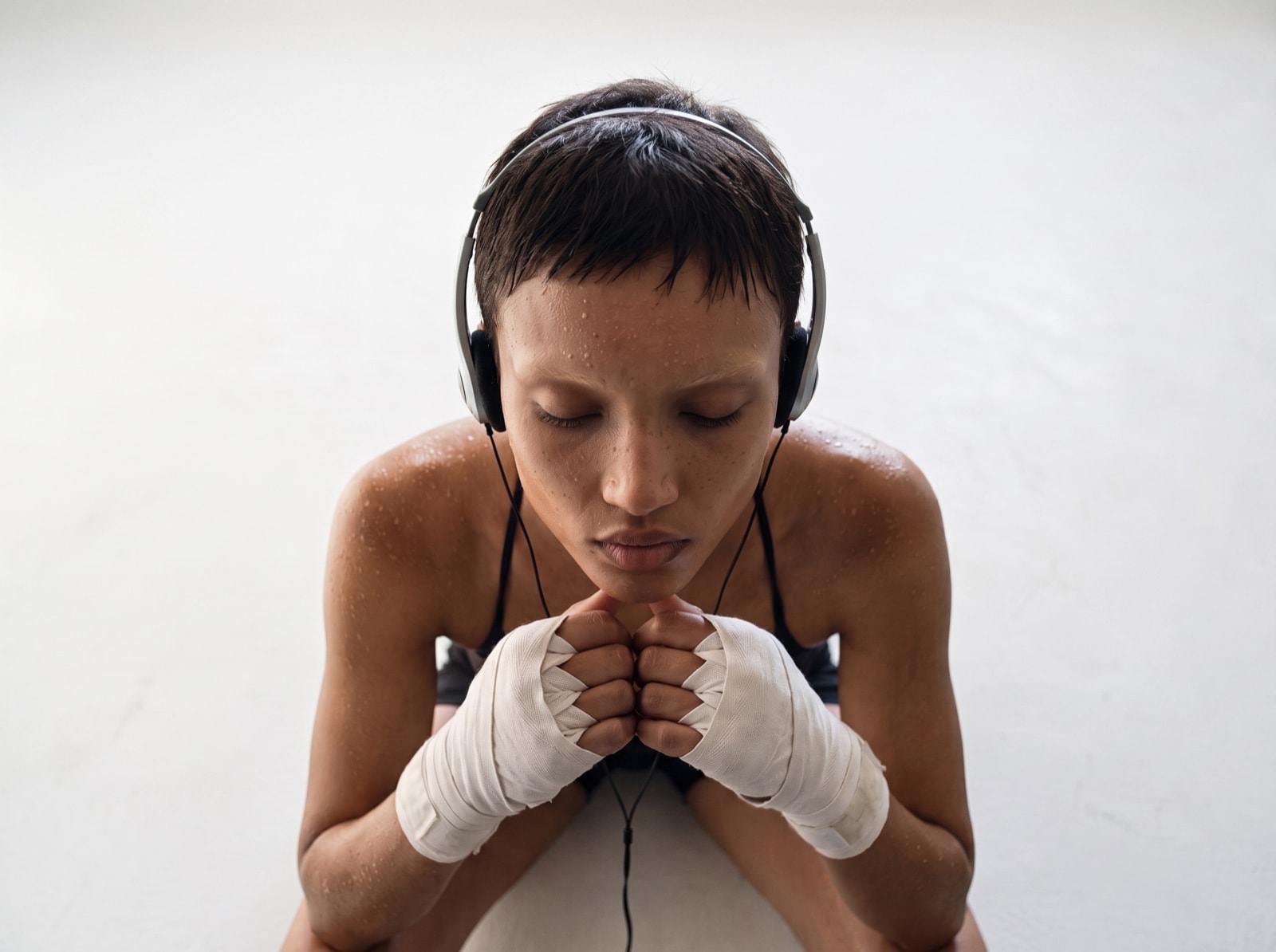 A boxer in headphones bows her head over wrapped fists, eyes closed, against a white wall.